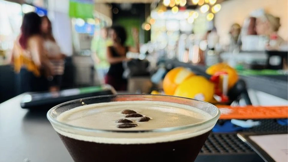 A close-up of a coffee cocktail with coffee beans on top, with blurred figures at a bar in the background.